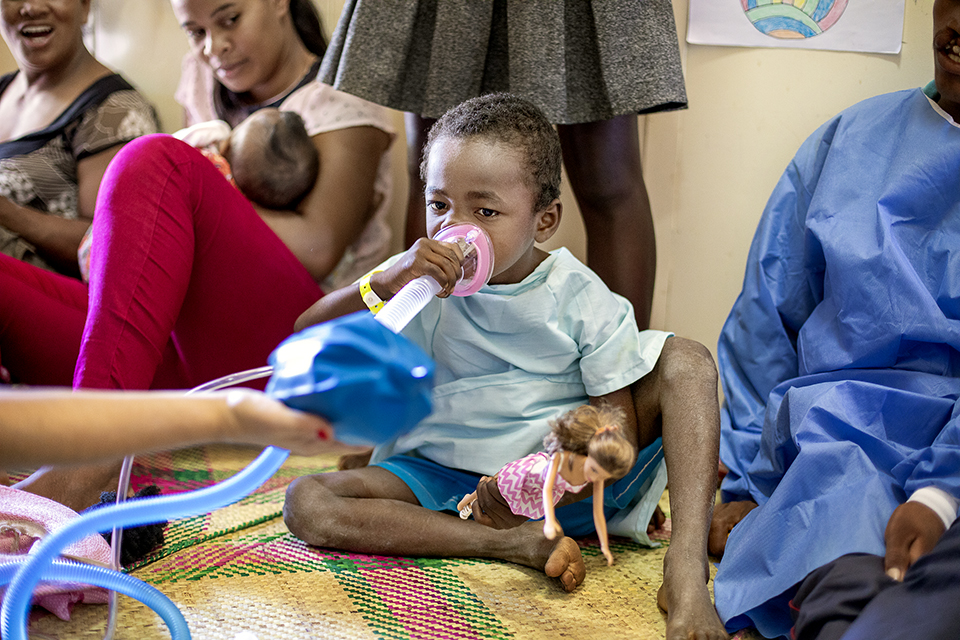 A young child sits on a mat holding a doll and wearing a hospital gown while using an inhaler mask. Other people, including women and children, sit around them indoors, some smiling, in a brightly lit room.