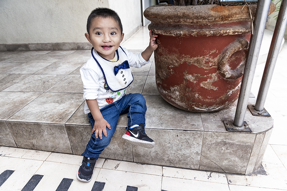 A smiling young boy in jeans, a white shirt with a bowtie print, and black Nike sneakers sits on a tiled step beside a large, weathered terra-cotta planter, holding onto its rim with one hand.
