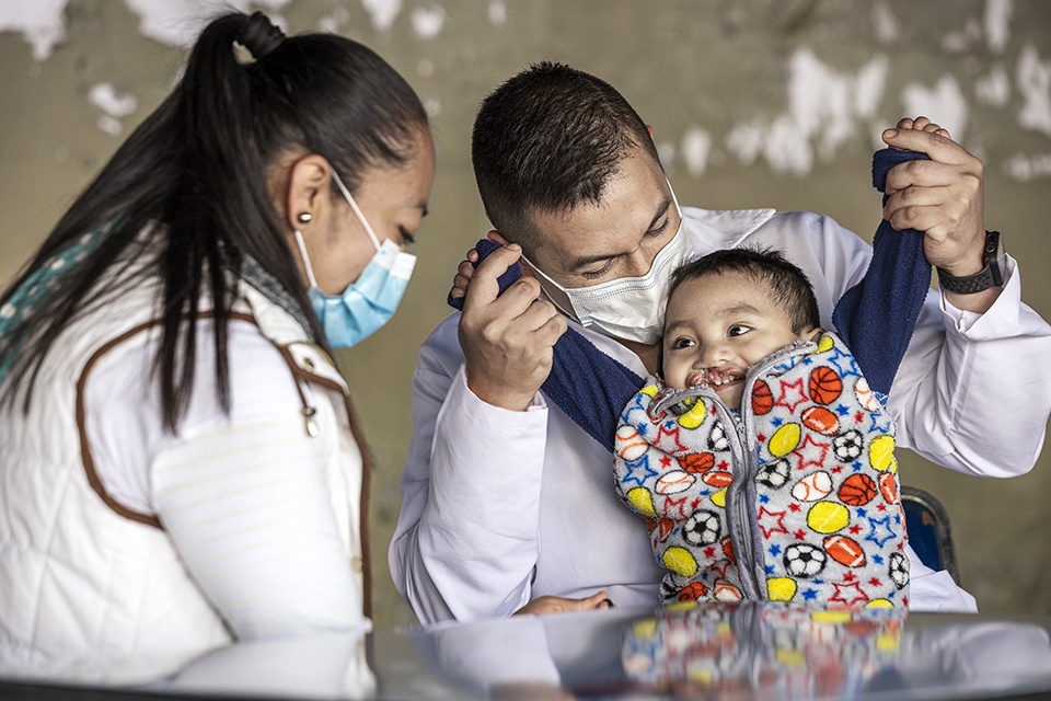 A smiling baby in a colorful jacket sits on an adult’s lap while another adult looks on. Both adults wear face masks and appear to be caregivers or medical workers. The scene is warm and caring.