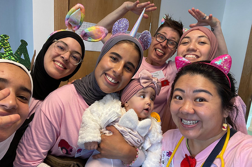 A group of smiling people wearing fun animal ear headbands and pink shirts pose for a selfie, with one woman holding a baby dressed in white. They appear happy and cheerful in a festive indoor setting.