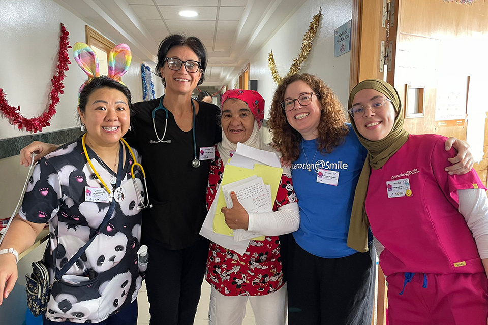 Five smiling healthcare workers stand together in a hallway decorated with tinsel. They wear colorful scrubs, badges, and stethoscopes; one has bunny ears and another holds papers, giving a thumbs-up.