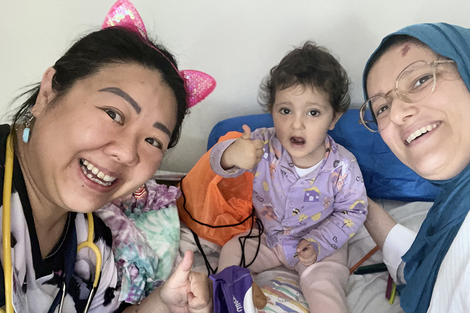 Two smiling women, one wearing cat ears and a stethoscope, and the other in glasses and a blue headscarf, sit with a young child who is giving a thumbs-up gesture. All appear happy and cheerful indoors.