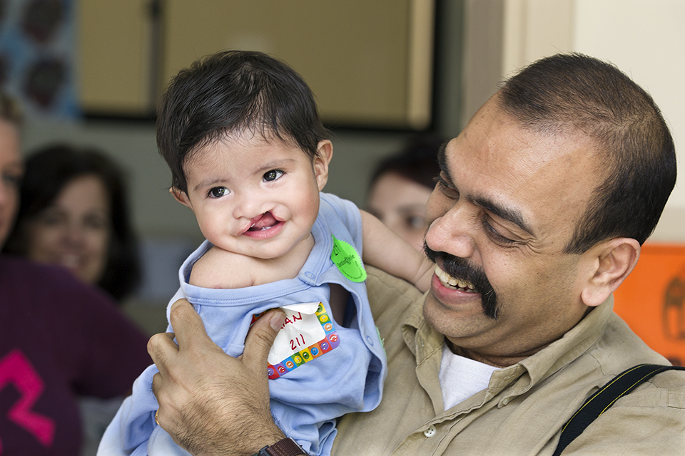 A smiling man holds a baby with a cleft lip, both appearing happy. The baby wears a light blue shirt, and there are other people blurred in the background.
