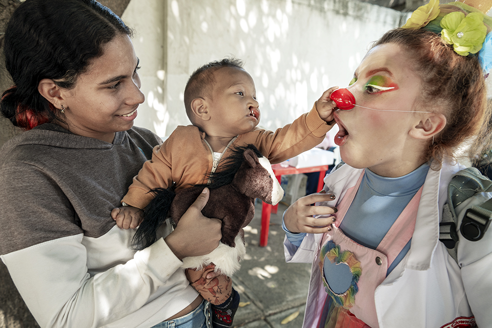 A woman holds a baby who reaches out to touch the red nose of a colorful clown. The clown smiles and leans in playfully, while the baby holds a stuffed toy horse. The scene takes place outdoors, shaded by trees.