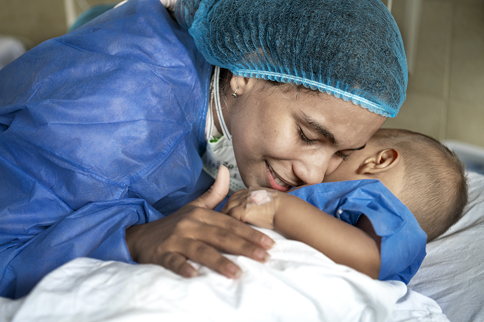 A woman in a blue surgical gown and hair cover lovingly hugs a small child in a hospital bed. Both appear emotional and close, sharing a tender moment in a medical setting.