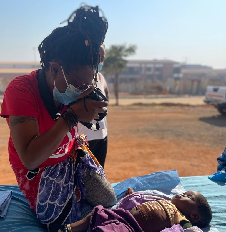 A woman wearing a mask and glasses takes a close-up photo of a young child lying on a bed outdoors, with buildings and a tree visible in the background.