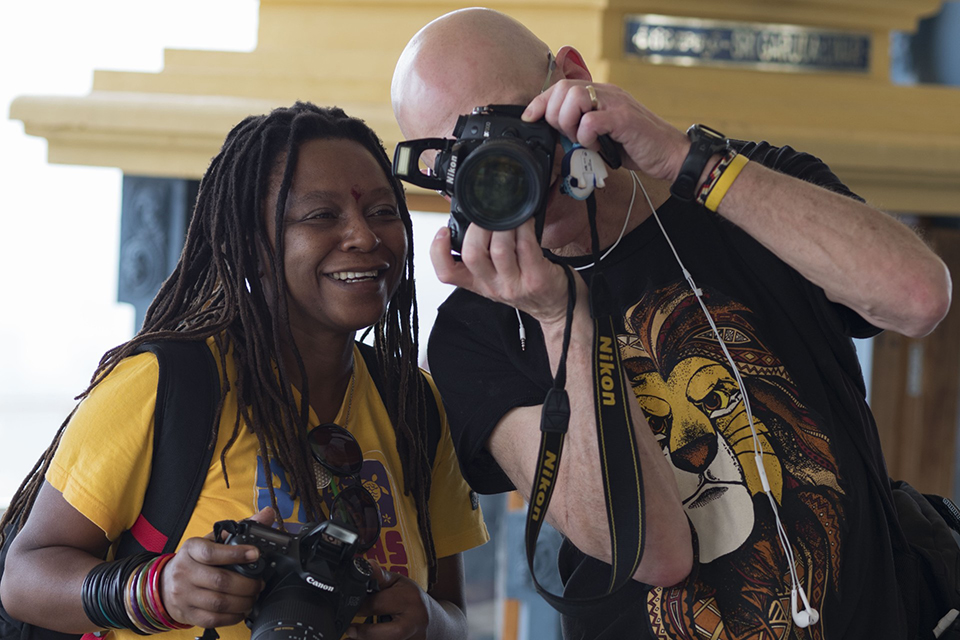 Two people, one with long dreadlocks and another bald, smile and look at the screen of a Nikon camera. Both hold cameras, appearing happy while photographing or reviewing photos together indoors.
