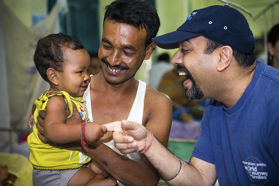 A smiling man in a blue shirt gently holds the hand of a cheerful baby while another man, likely the father, holds the baby and smiles warmly. The scene is joyful and caring, set indoors.