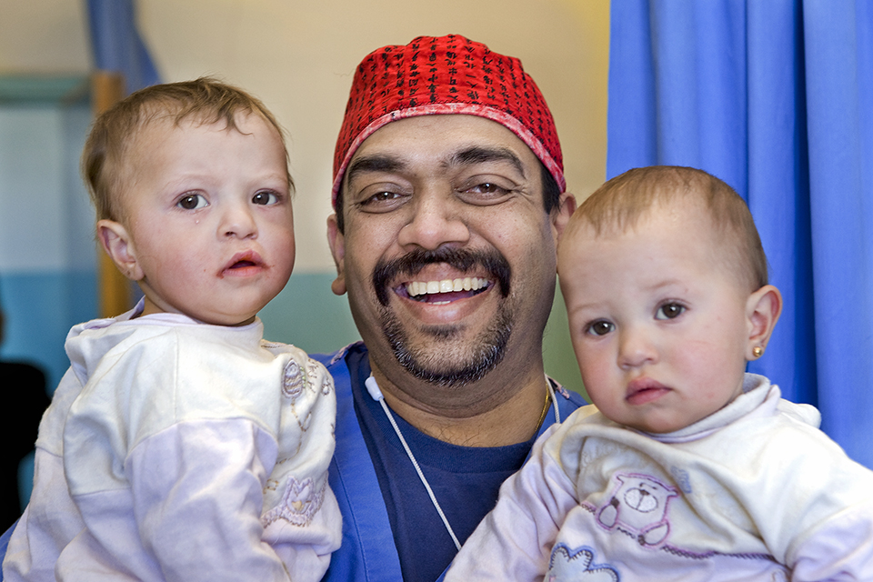 A smiling man in a red surgical cap holds two young children in white shirts, one on each arm. The children have short hair and neutral expressions. Blue curtains and a doorway are visible in the background.