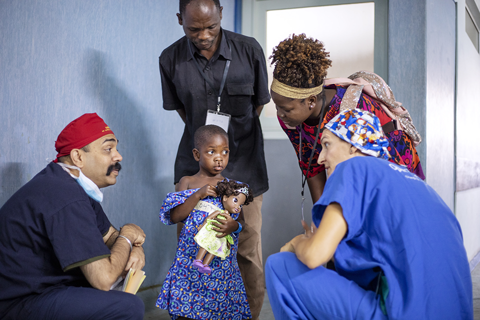 Two doctors in scrubs crouch to speak with a young girl holding a doll, while two adults stand behind her. All appear engaged in conversation in a hospital setting.
