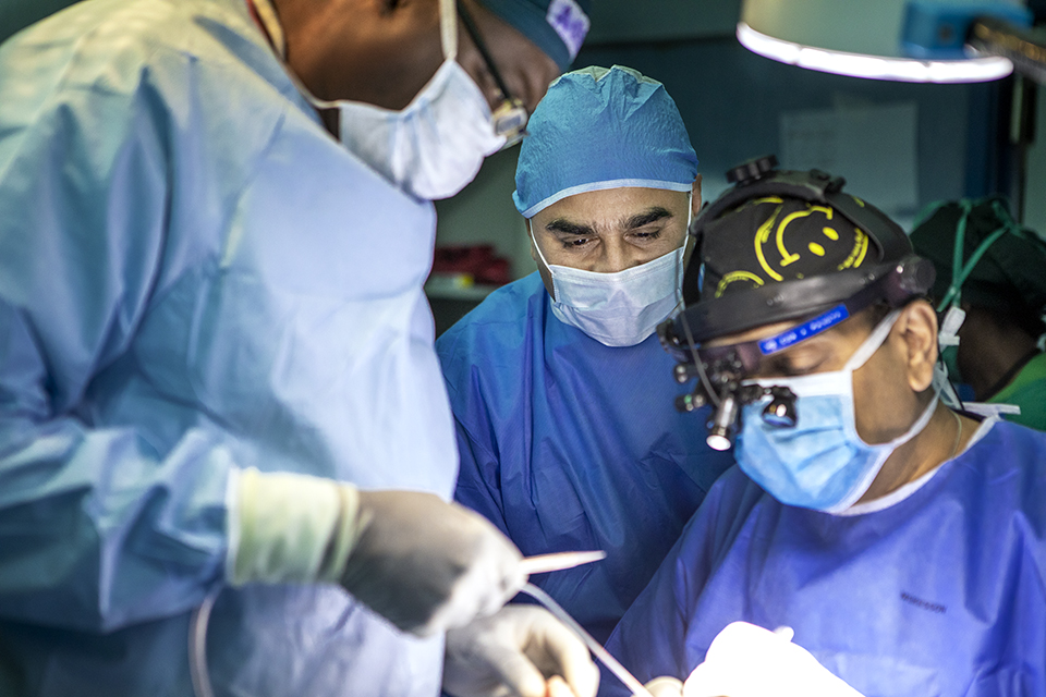 Three surgeons in blue scrubs and masks perform a procedure in an operating room, with one using surgical tools and another wearing a headlamp for focused lighting.