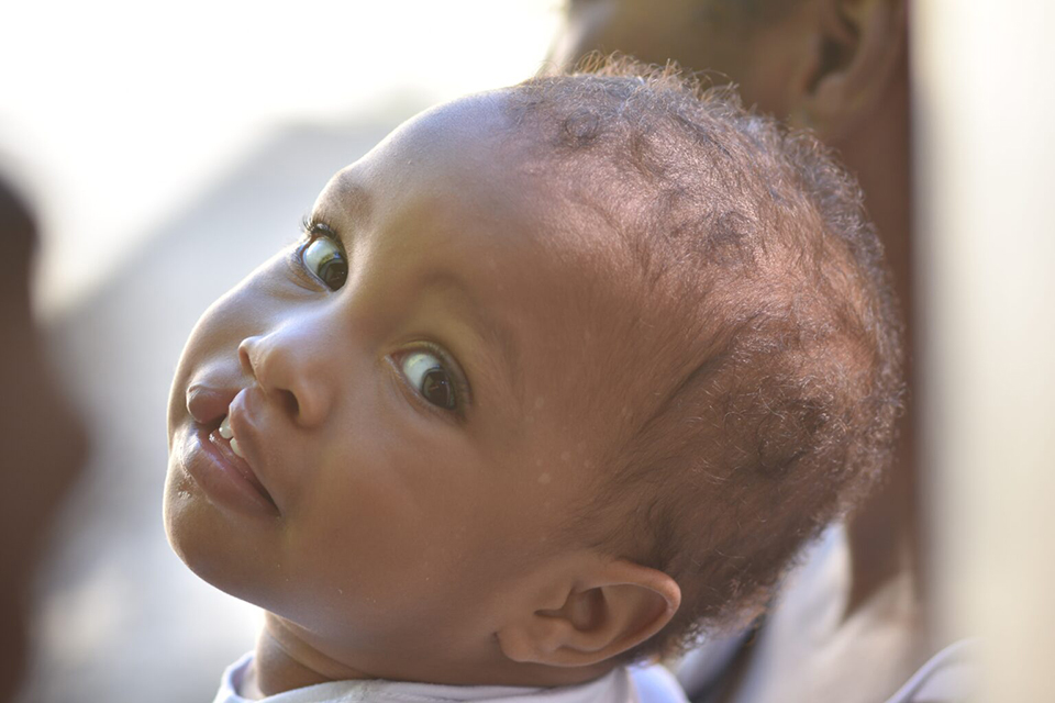 A baby with curly hair looks over their shoulder, gazing softly at the camera with wide eyes and a slight smile, while sunlight gently illuminates their face.