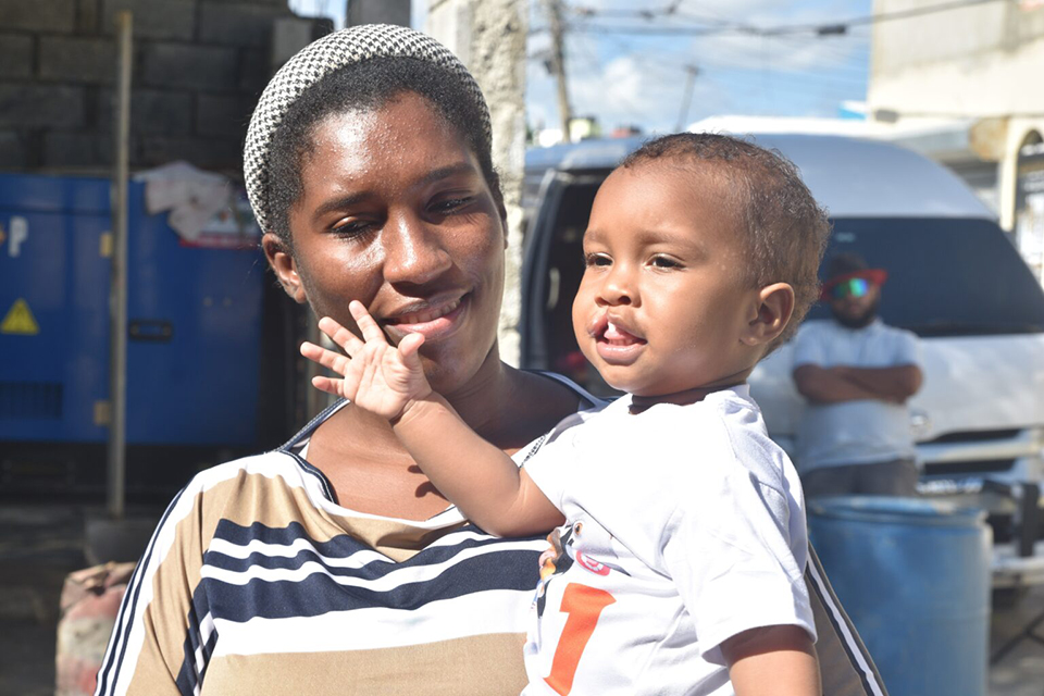 A woman smiles while holding a baby who is waving. They are outdoors on a sunny day, with a van and another person blurred in the background.