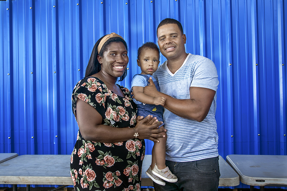 A smiling woman in a floral dress stands beside a man in a striped shirt holding a toddler. They pose in front of a bright blue corrugated metal wall, with tables in the foreground.