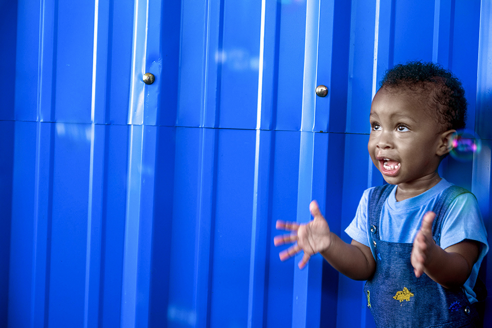 A young child in denim overalls excitedly reaching out with both hands, standing in front of a bright blue corrugated metal wall. The child looks delighted, playing with bubbles.