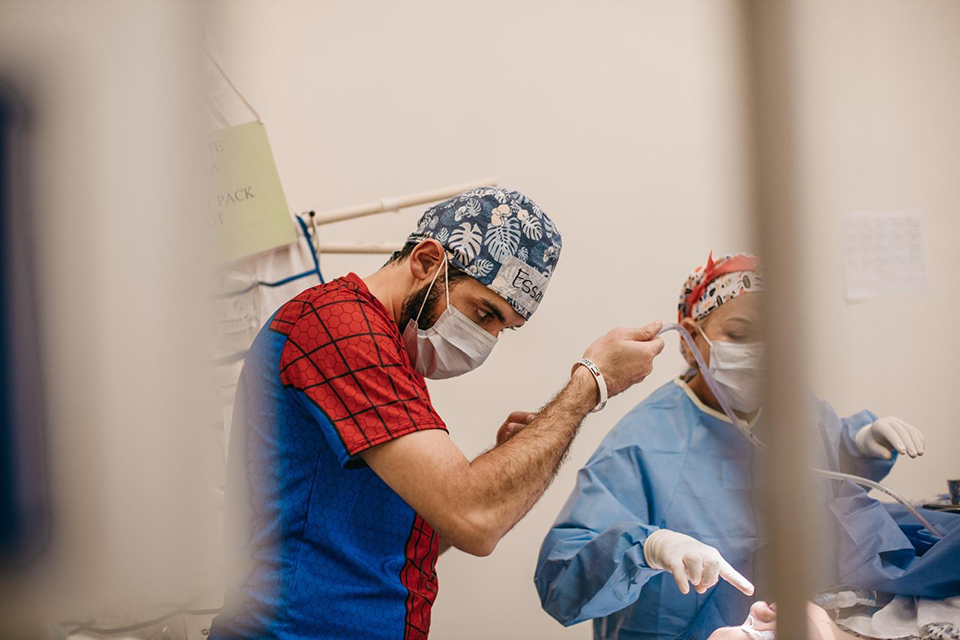 An anaesthetist in a blue and red Spiderman-themed scrub top and cap works alongside a masked nurse in blue surgical attire during an operation in a brightly lit medical room.