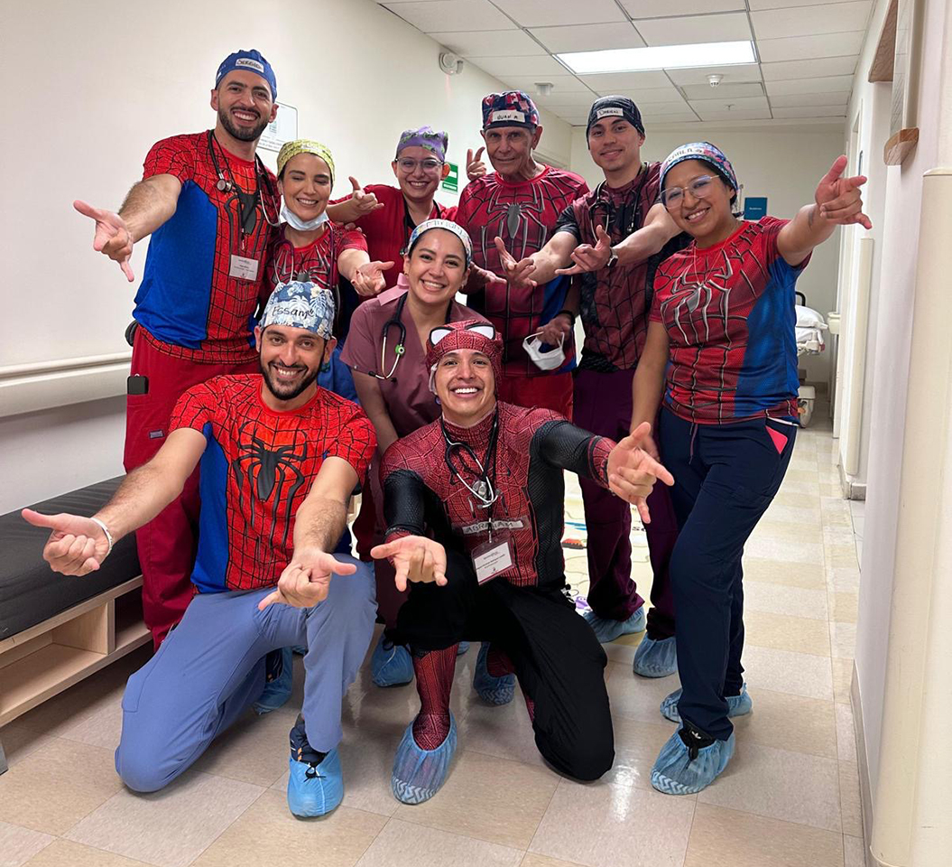 A group of nine smiling healthcare workers in medical scrubs and shoe covers pose together in a hallway, wearing Spider-Man shirts and costumes, playfully reaching their hands forward like Spider-Man shooting webs.