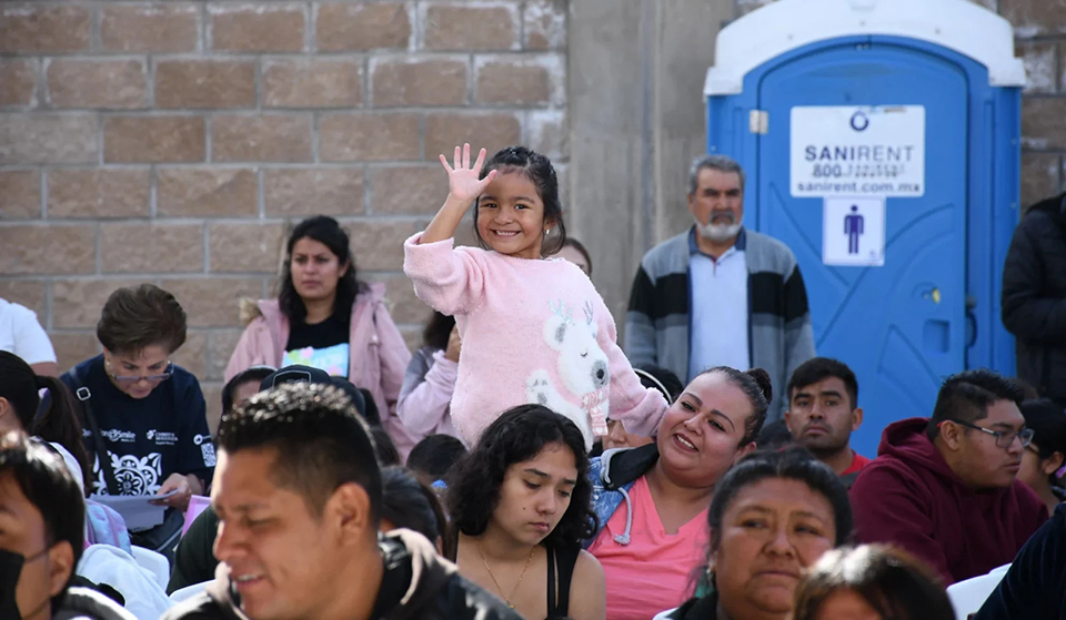 A young girl in a pink sweater smiles and waves while sitting on a woman's lap in a crowd. People around her are seated outdoors in front of a stone wall.