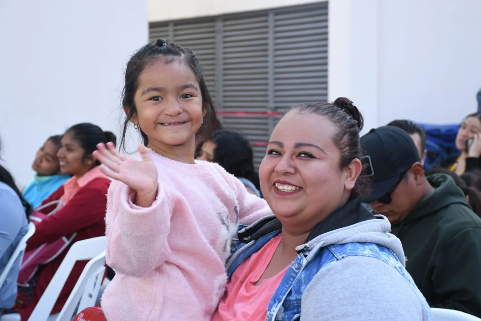 A young girl in a pink sweater smiles and waves while sitting on a smiling woman’s lap. They are surrounded by other people seated outdoors, all appearing cheerful and relaxed.