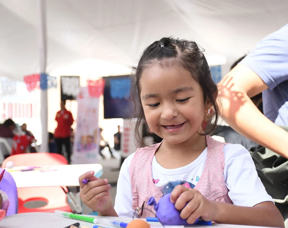 A young girl smiles while crafting at a table, holding a purple object. Art supplies are spread out in front of her. The background shows other people and colorful decorations in a bright, tented space.