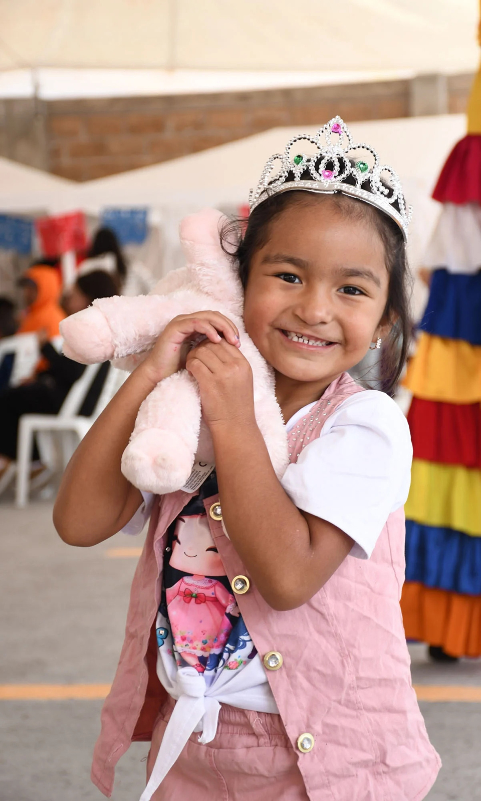 A young girl wearing a tiara and pink vest smiles while hugging a plush teddy bear. She stands indoors, with colorful decorations and people in the blurred background.