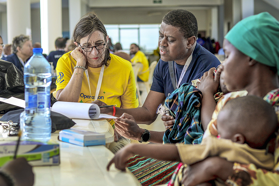 A woman in a yellow shirt and a man speak with a woman holding a child at a table. Papers, a water bottle, and medical supplies are on the table, with other people gathered in the background.