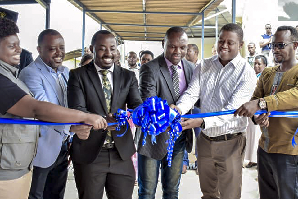A group of six people smile as they hold and cut a blue ribbon with a large bow, during what appears to be a ribbon-cutting ceremony. Others are gathered behind them, watching the event.