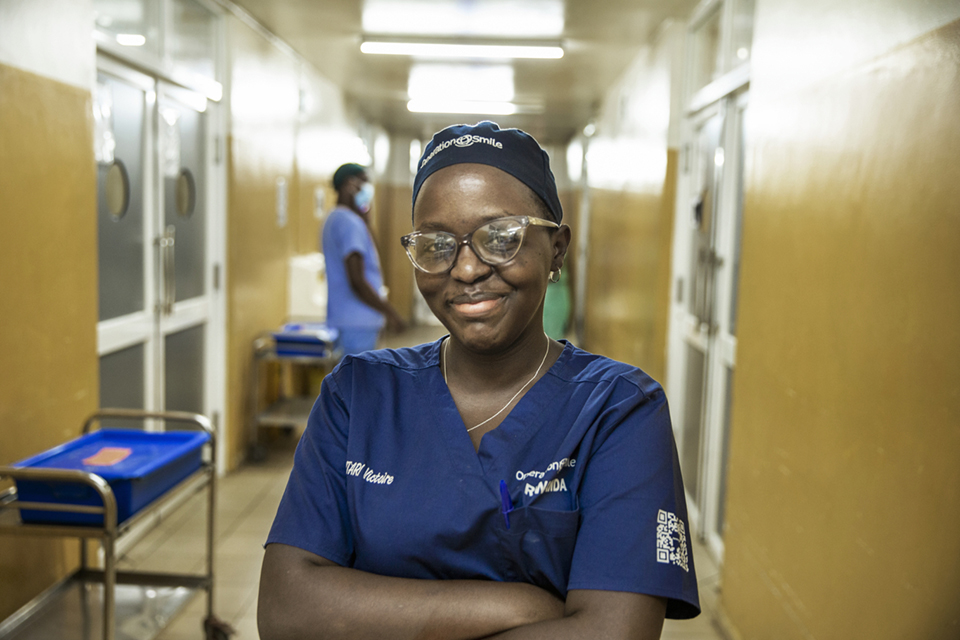 A healthcare worker wearing blue scrubs, glasses, and a head covering stands smiling in a hospital hallway with her arms crossed. Other medical staff and equipment are visible in the background.