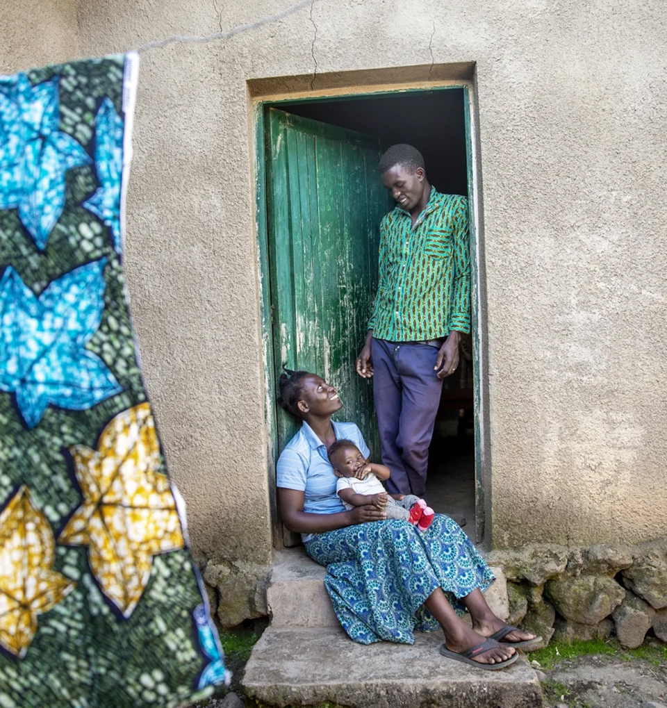 A woman sits on a doorstep, holding a baby and smiling up at a man standing in the doorway. The building wall is brown, the door is green, and a colorful fabric hangs in the foreground.