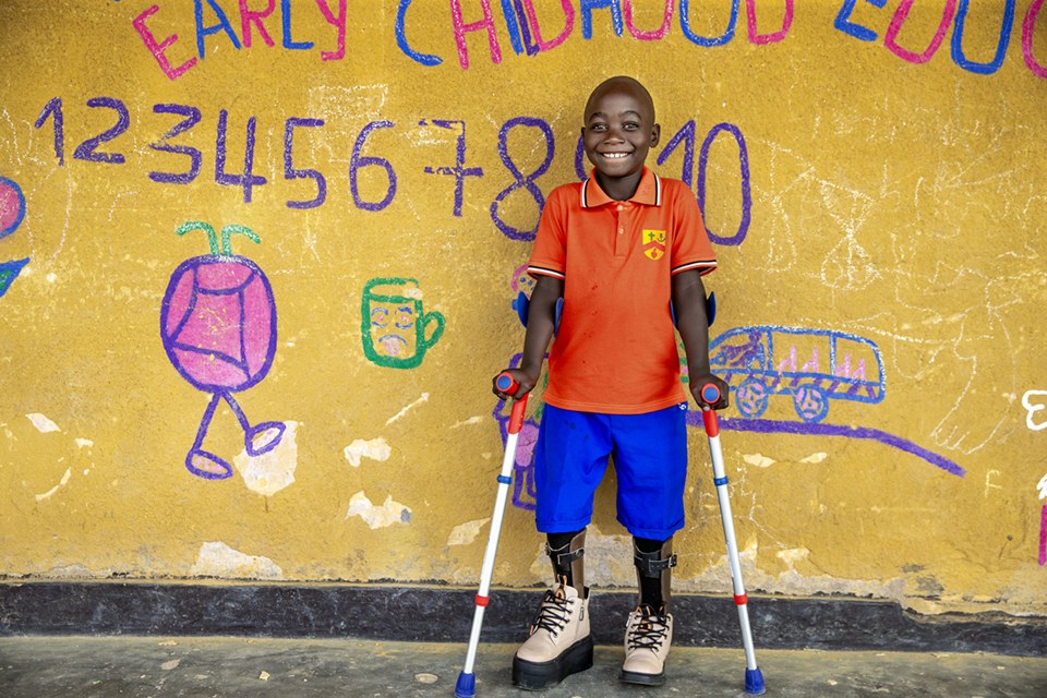 A smiling boy with prosthetic legs and forearm crutches stands in front of a yellow wall decorated with colorful drawings and numbers at a school.