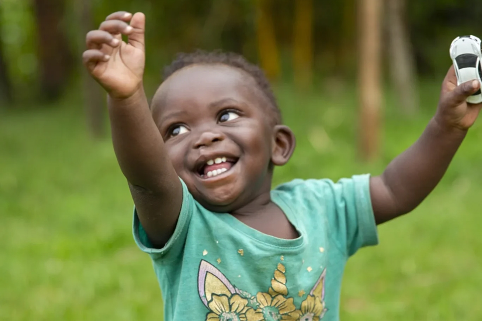 A smiling toddler in a green unicorn shirt raises one arm joyfully while holding a toy car in the other hand, standing outdoors on green grass.