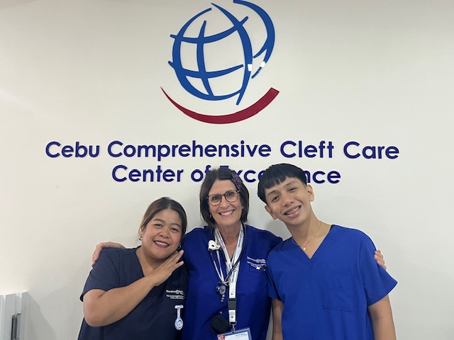 Three smiling healthcare workers stand in front of a wall sign that reads, "Cebu Comprehensive Cleft Care Center of Excellence.” Two wear dark blue scrubs and one wears a navy shirt with a lanyard and stethoscope.